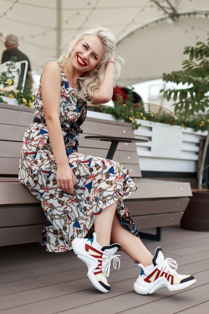 Feliz linda garota em um vestido vintage com padrão e tênis da moda, sentado e relaxando em um banco na cidade