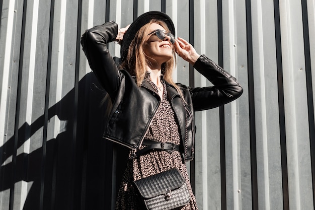 Feliz linda garota na moda com óculos de sol estilizados em um vestido vintage com jaqueta de couro preta e bolsa perto de uma parede de metal