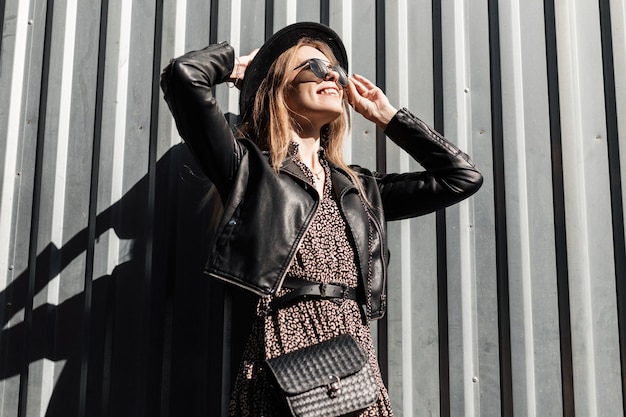 Foto feliz linda garota na moda com óculos de sol estilizados em um vestido vintage com jaqueta de couro preta e bolsa perto de uma parede de metal