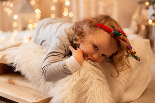 Feliz linda garotinha criança de pijama encontra-se na cama no contexto das luzes de natal e decorações.