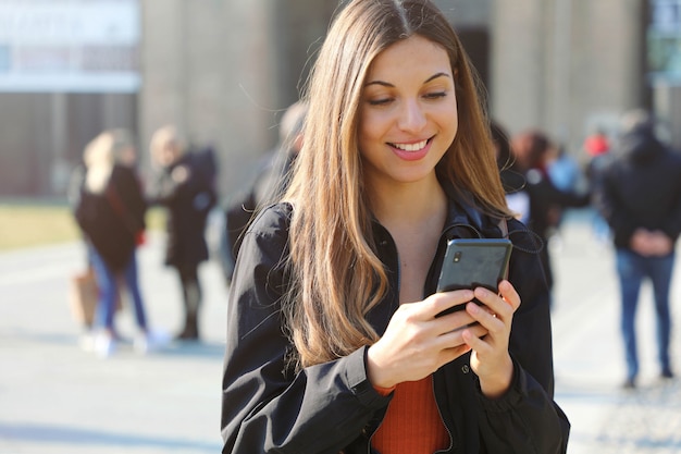 Feliz linda jovem colegial com telefone inteligente ao ar livre em um dia ensolarado de primavera, enviando mensagens de texto e sorrindo