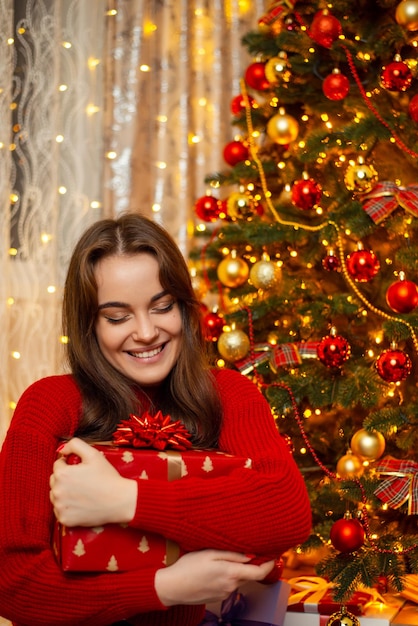 Feliz linda jovem de suéter vermelho com cabelo castanho abraça uma grande caixa de presente árvore de Natal e luzes de guirlanda no fundo