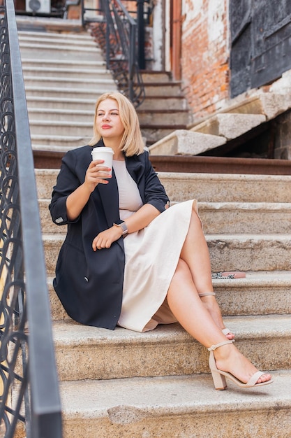 Feliz linda jovem de vestido e jaqueta anda pela cidade e bebe café