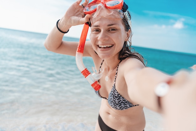 Foto feliz linda jovem encantadora com um lindo sorriso de biquíni em uma máscara para mergulho leva uma selfie à beira-mar, atividades ao ar livre, atividades de praia