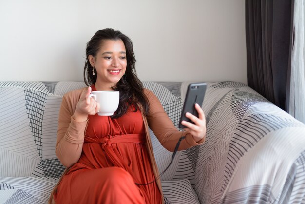 Feliz linda jovem mulher indiana tomando café enquanto estiver a tomar selfie em casa