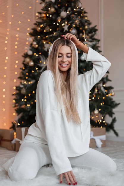 Feliz linda jovem sorridente em uma roupa branca de moda com capuz sentado no chão da casa perto da árvore de Natal