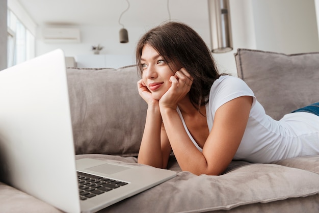 Feliz linda joven tumbado en el sofá y usando la computadora portátil en casa