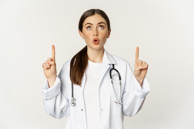 Feliz linda médica médica apontando os dedos para cima mostrando propaganda em cima de branco...