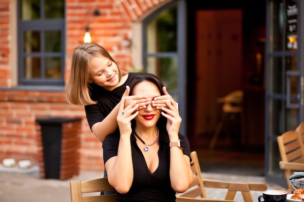 Feliz linda menina adorável está cobrindo os olhos da mãe com as palmas das mãos.