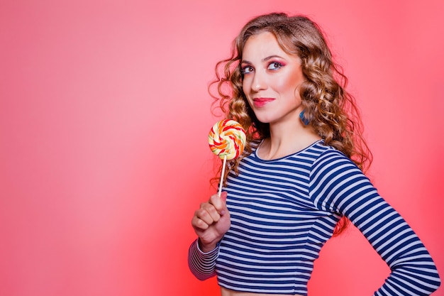 Feliz linda menina morena com maquiagem vermelha, cabelo encaracolado e um suéter de listras azuis posando contra um fundo vermelho. Foto horizontal