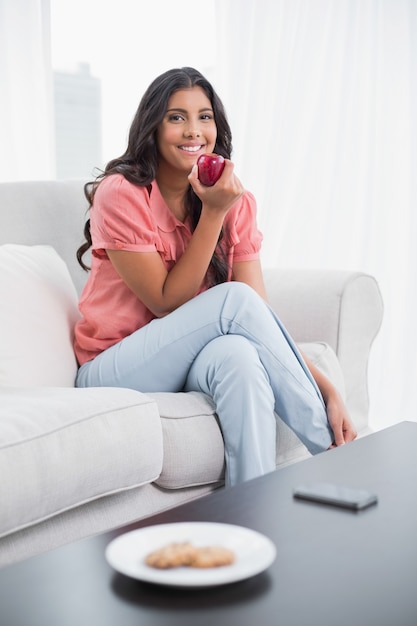 Feliz linda morena sentada en el sofá con manzana roja