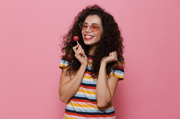 Feliz linda mujer joven posando aislada en rosa come piruleta de caramelo.