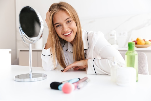 Foto feliz linda mujer en pijama haciendo maquillaje y mirando a la cámara mientras está sentado a la mesa en una habitación luminosa