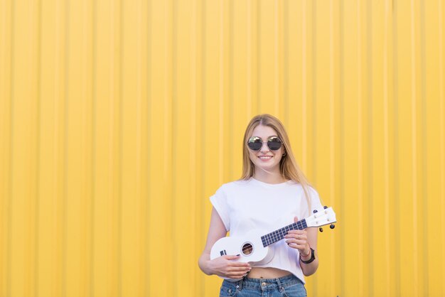 Feliz, linda mujer toca el ukelele contra la pared amarilla y sonríe. Concepto musical