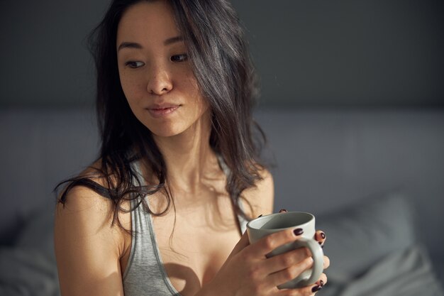 Feliz linda mulher asiática segurando uma xícara e saboreando seu café da manhã