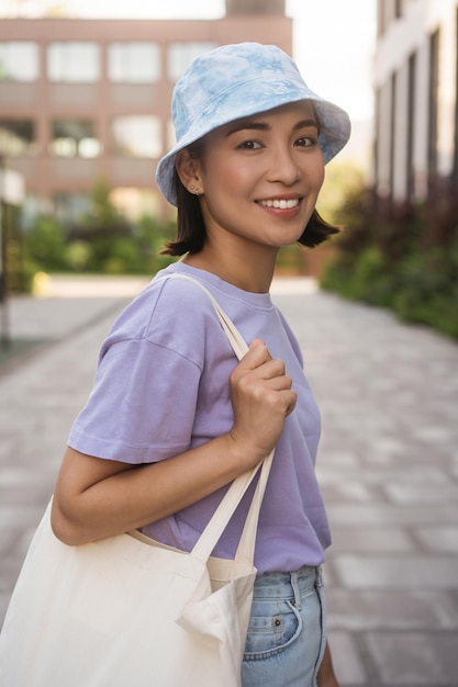 Feliz linda mulher asiática vestindo roupas casuais, panamá estiloso, segurando sacola de compras