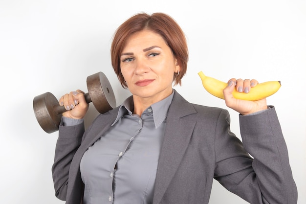 Foto feliz linda mulher de negócios de terno levanta um haltere e uma banana nas mãos. fitness e saúde. nutrição adequada e excelentes resultados.