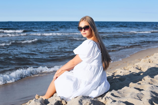 Feliz, linda mulher loira está sentada na praia do oceano em um vestido branco de verão e óculos de sol.