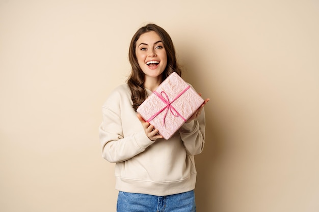 Feliz linda namorada segurando a caixa de presentes rosa e rindo, sorrindo alegremente, conceito de feriados e celebração, fundo bege