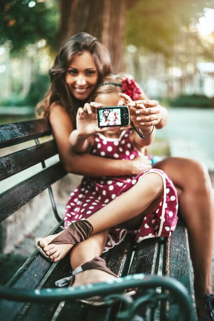 Feliz linda niña y su madre están posando para un autorretrato en un banco en el parque.