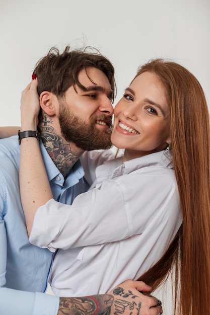 Feliz lindo casal elegante homem hipster bonito e mulher ruiva bonita com um sorriso em uma camisa da moda abraçam no estúdio em um fundo branco