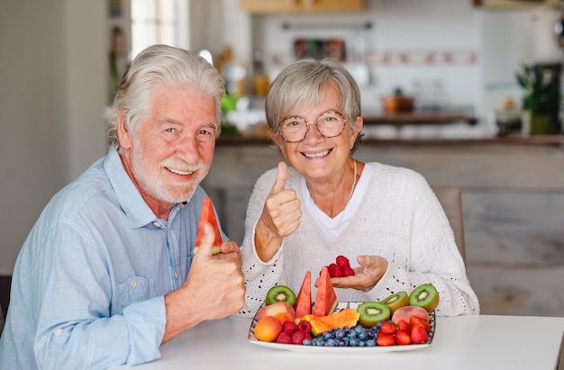 Foto feliz lindo casal sênior aposentado tendo uma pausa com frutas frescas da estação em casa polegar para cima olhando para o conceito de alimentação saudável da câmera