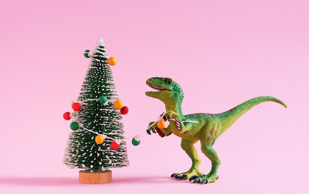 Feliz lindo dinosaurio verde decora el árbol de Navidad con guirnalda de bombillas retro Tarjeta de felicitación de Navidad y año nuevo sobre fondo rosa pastel