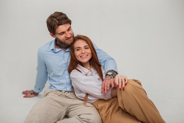 Feliz lindo jovem casal de amantes em roupas da moda estão sentados no estúdio em um fundo branco Linda garota ruiva feliz com sorriso fofo e homem bonito hipster