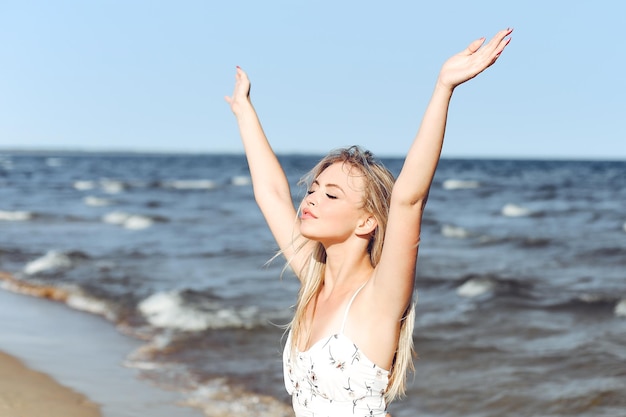 Feliz loira bela mulher na praia do oceano de pé em um vestido de verão branco, levantando as mãos.