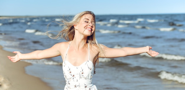 Feliz loira bela mulher na praia do oceano de pé em um vestido de verão branco, levantando as mãos.