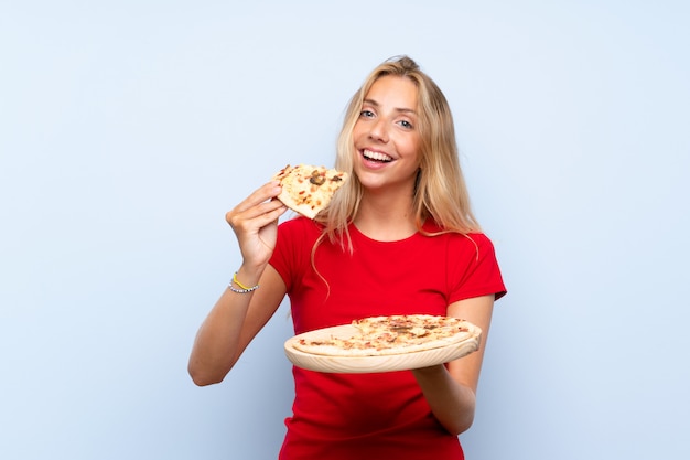 Feliz loira jovem segurando uma pizza sobre parede azul isolada