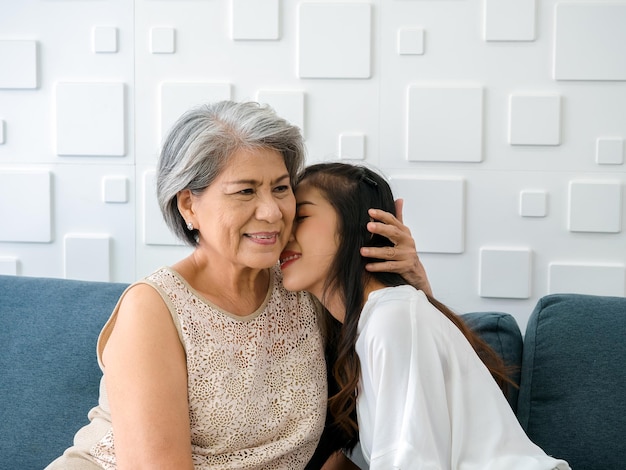 Feliz madre asiática de cabello blanco abrazando a su hermosa hija mientras besa su mejilla con cariño y sonríe mientras se sienta en un sofá con fondo blanco en la sala de estar en casa