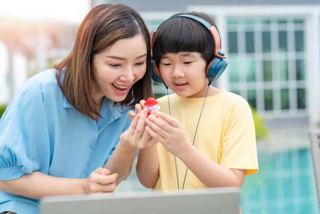 Feliz madre asiática le da un juguete a su hijo mientras el niño usa auriculares con una laptop en casa