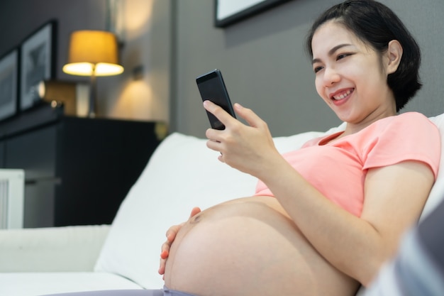 Feliz madre asiática sonriente mediante teléfono móvil durante el embarazo en casa.