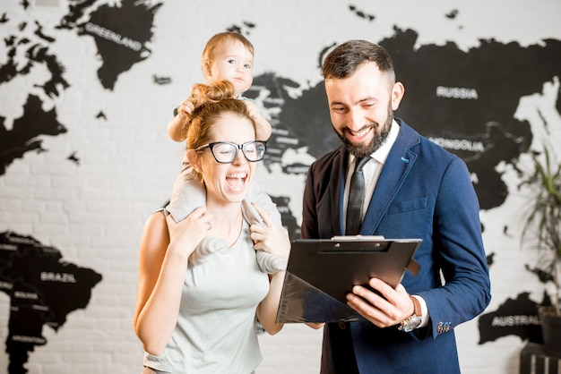 Feliz madre y bebé eligiendo un tour para unas vacaciones de verano con un agente masculino en la oficina de la agencia de viajes con un mapa del mundo en el fondo