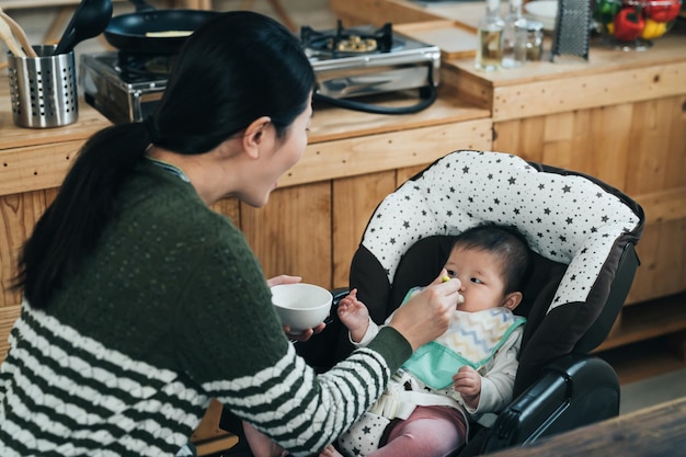 feliz madre coreana sosteniendo un arco y una cuchara está alimentando al bebé a la hora del almuerzo. una madre asiática amorosa está diciendo "ah" mientras le da a su hijo comida sólida en casa.