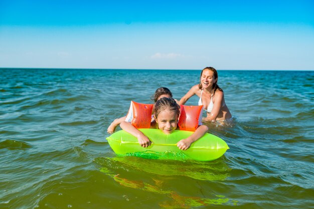 Feliz madre y dos pequeñas hijas positivas se bañan y nadan en el mar con un colchón de aire en un día soleado de verano