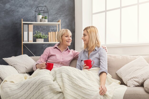 Feliz madre e hija adultas cubiertas con una alfombra pasando tiempo juntas, charlando, compartiendo noticias y tomando café en casa el fin de semana. Concepto de generación y relación