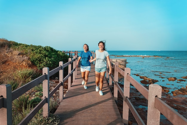 Feliz madre e hija corriendo en la acera junto al mar. Sonriente mujer de mediana edad y adolescente cogidos de la mano y caminando al atardecer, tiro de longitud completa. Maqueta de camiseta