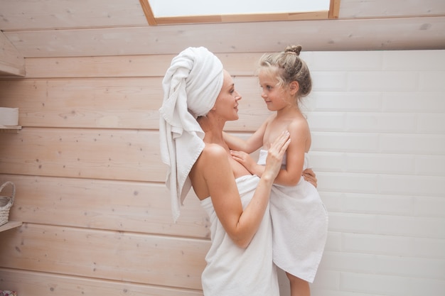 Feliz madre e hija de familia amorosa con toallas pasar tiempo juntos en el baño blanco.