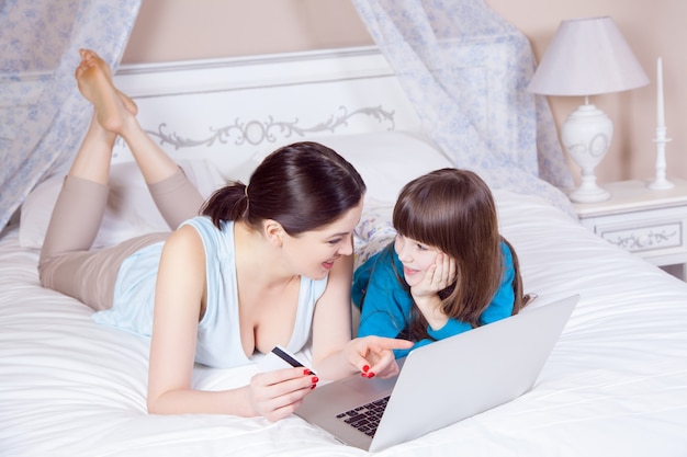 Feliz madre e hija hacen compras en línea y pagan con tarjeta de crédito con el portátil acostado en la cama en el dormitorio. Familia feliz. Tiro del estudio.