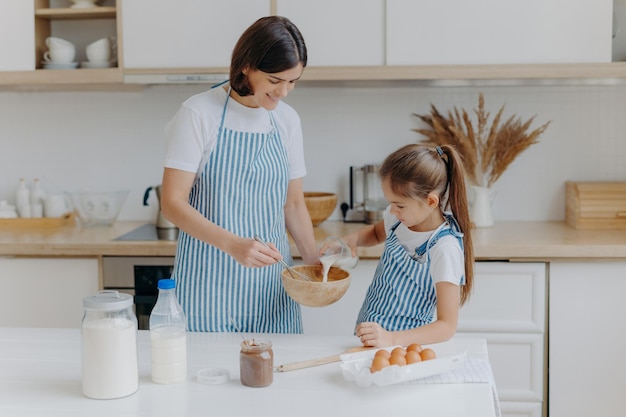 Feliz madre e hija hornean juntas en la cocina usan diferentes ingredientes usan delantales se paran contra el interior de la cocina la niña vierte leche en el arco Mamá cariñosa enseña al niño a cocinar o hacer masa