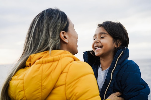 Foto feliz madre e hija latinas divirtiéndose juntas en la playa durante el invierno centrarse en la cara de la niña