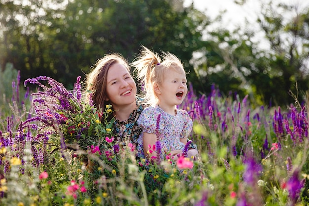 Feliz madre e hija rubia en un campo floreciente de verano con un ramo de flores silvestres en sus manos