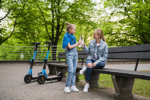 Feliz madre e hija con scooters eléctricos beben limonada en el parque de la ciudad