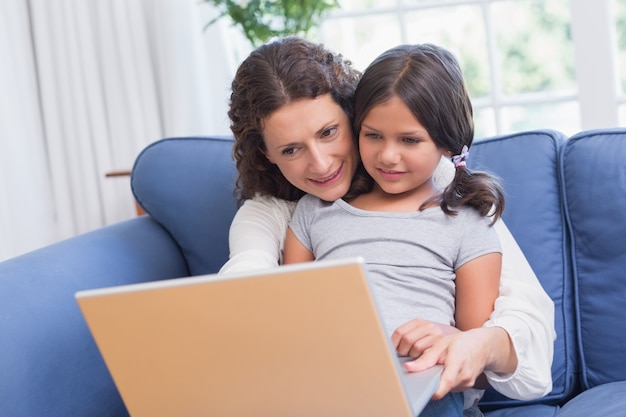 Feliz madre e hija sentada en el sofá y usando la computadora portátil