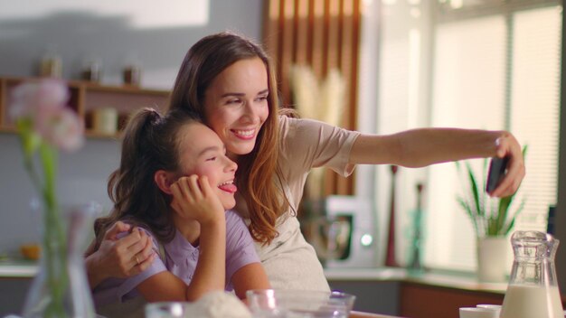 Foto feliz madre e hija tomando una foto selfie en un teléfono inteligente en la cocina doméstica niña y mujer sonrientes usando un teléfono celular para autorretrato juntas en casa en cámara lenta