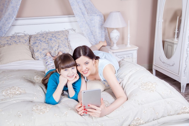 Feliz madre e hija yacían en la cama con tableta digital, sonriendo y mirando a cámara. Tiro del estudio.