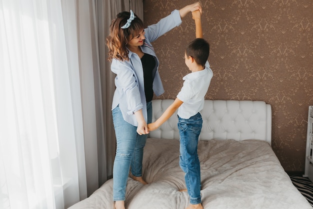 Foto feliz madre e hijo están saltando en la cama, tomados de la mano, riendo mamá está jugando con el niño en el dormitorio.