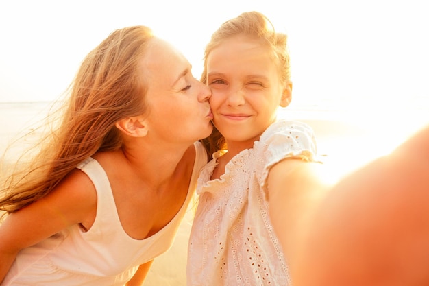 Feliz madre con estilo besa a su hija y se toma una selfie en la playa de arena en una puesta de sol. Día de la madre. Niña rubia y hermosa mujer tomando fotos por teléfono. Turismo en el extranjero. Videollamadas en línea.